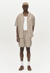 Camicia beige a maniche corte, canottiera bianca, pantaloni corti beige, scarpe marroni e cappellino beige con il logo "Wilderness". Tessuto morbido, vestibilità rilassata.