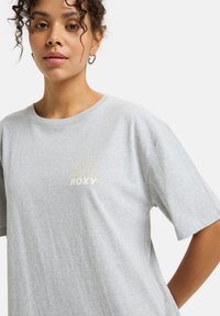 Szara bawełniana koszulka z luźnym krojem, okrągłym dekoltem i krótkimi rękawami. Posiada złote logo "90 Roxy" na piersi.