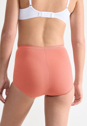 Persona vista da dietro che indossa un reggiseno bianco e mutandine a vita alta color corallo su uno sfondo chiaro e uniforme.