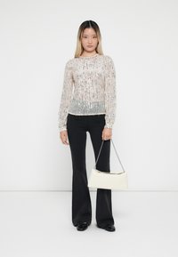 Top long transparent à manches longues avec des sequins argentés, superposé sur une section grise unie, associé à un pantalon noir évasé et un sac blanc avec une chaîne.