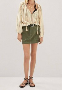 Lys beige, stribet bluse med løs pasform og snore, parret med en grøn denimnederdel. Accentueret med et sort bælte og nitter sandaler.