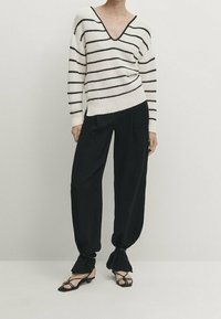 Pull crème tricoté à rayures horizontales noires, col en V plongeant et ourlet côtelé, associé à un pantalon noir ample, stylisé avec des chaussures noires à lanières.