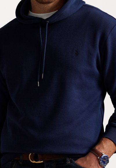 Hoodie azul-marinho com um tecido texturizado, apresentando um bolso canguru e cordões. Pequeno logótipo no peito. Combinado com um cinto e um relógio castanho.