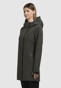 Giacca impermeabile verde con cappuccio, maniche lunghe, tasche laterali con zip e taglio dritto, dotata di cordini regolabili sul cappuccio.