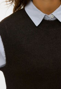 Donkerbruin gebreid vest over een lichtblauw overhemd met kraag, voorzien van een ronde hals en een zachte textuur, geschikt voor layering.