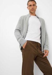 Un sweat à zip gris clair avec des poignets côtelés, porté sur un t-shirt blanc et associé à un pantalon marron, présenté sur un fond blanc.