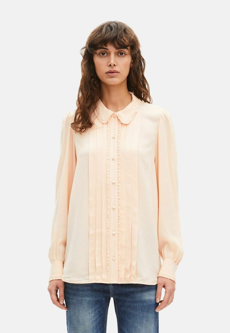 Mujer con cabello castaño ondulado, vistiendo una blusa de manga larga color durazno pálido con volantes y botones, combinada con jeans azules, de pie frente a un fondo blanco.