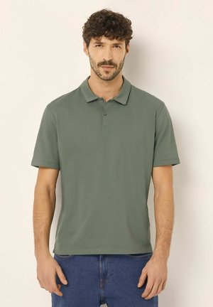 Dunkelgrünes Poloshirt mit Kragen, kurzen Ärmeln und einer dreiknöpfigen Knopfleiste. Glatte Stoffstruktur, kombiniert mit blauen Jeansshorts.