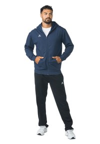 Marineblauwe hoodie met rits, voorzakken en capuchon, gecombineerd met een zwarte broek. Beide hebben merkaccenten in het wit. Lichtgewicht stof.