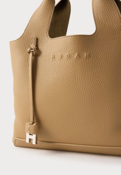 Ręczna torba typu tote w kolorze jasnobrązowym ze strukturą ziarnistą, wyposażona w wycięte uchwyty oraz odpinany pasek z metalicznym akcentem. Logo wytłoczone z boku.