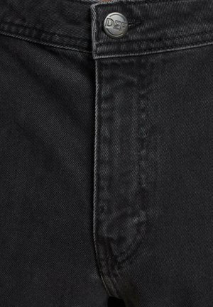Jean en denim noir avec une fermeture éclair/bouton, présentant des coutures contrastées et un bouton en métal brandé à la taille.