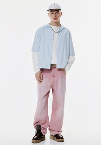Chemise à manches courtes rayée bleu clair sur une chemise à manches longues blanche, associée à un jean baggy rose dégradé et des bottes noires à lacets.