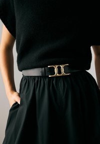 Ceinture en cuir noir avec une boucle double cercle en ton doré. Associée à un haut noir et une jupe, mettant en valeur le design de la boucle.
