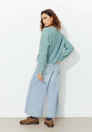 Pull en maille vert menthe, coupe loose ; jeans bleu clair à jambes larges ; chaussures léopard marron avec accents noirs ; arrière-plan de studio.