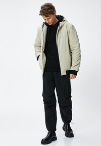 Lichtgrijze hoodie-jack over een zwart sweatshirt, gecombineerd met zwarte cargobroek en zwarte laarzen. Glad materiaal en een ontspannen pasvorm.