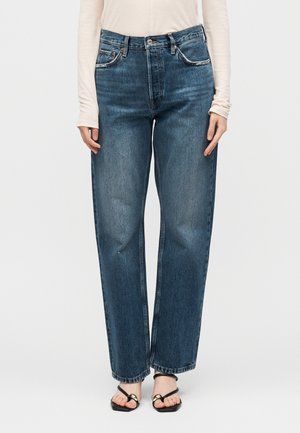 Kvinde iført blå jeans med høj talje og lige ben, lysebeige bluse med lange ærmer og sorte åbne sandaler med guldlukninger.