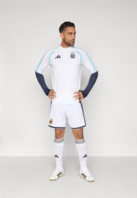 Άνδρας με άσπρη και ανοιχτό μπλε στολή ποδοσφαίρου της Αργεντινής στέκεται με τα χέρια στη μέση μπροστά σε διάφορο φόντο.