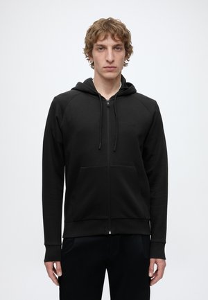 Homme aux cheveux châtain clair bouclés portant un sweat à capuche noir zippé avec des poches sur le devant et un petit logo "BOSS" sur le côté gauche de la poitrine.