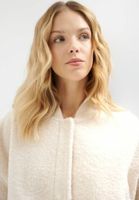 Jasno beżowy, teksturowany wełniany płaszcz z zaokrąglonym kołnierzem i pojedynczym zapięciem na guzik, prezentujący miękkie, puszyste wykończenie.