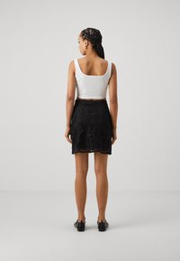 Hvit ermeløs crop topp og svart blonde miniskjørt med blomsterutskjæringer, kombinert med svarte sko. Bakside mot en ensfarget bakgrunn.