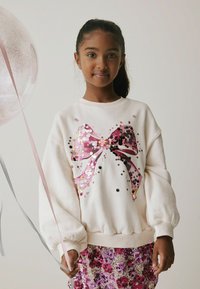 Hvid sweatshirt med et pink og sort pailletbue-design, ballon, og en skinnende pink og lilla pailletnederdel med puffede ærmer.
