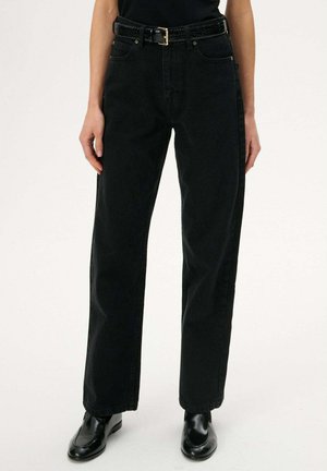 Osoba ubrana w czarne, proste jeansy z wysokim stanem, czarny pasek i czarne błyszczące mokasyny, stojąca na białym tle.