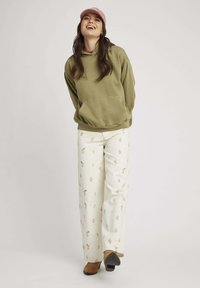 Hoodie olive avec une poche avant, associé à un pantalon large crème comportant un motif floral subtil. Des chaussures beige et grises complètent la tenue.