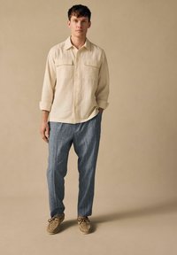 Beige strukturiertes Hemd mit zwei Brusttaschen, kombiniert mit blauen gestreiften Hosen und hellbraunen gewebten Schuhen. Schlichter Hintergrund.