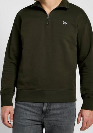 Homme portant un sweat-shirt vert foncé à fermeture éclair partielle avec un patch logo Lee sur la poitrine et un jean noir délavé, debout les bras détendus.