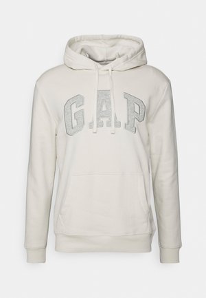 Kremowa bluza z kapturem z przednią kieszenią, regulowanym sznurkiem kaptura i dużymi szarymi literami "GAP" na piersi.