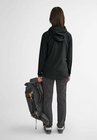 Veste à capuche légère noire, pantalon gris, tenant un sac à dos gris foncé avec des bretelles réglables et une finition texturée.