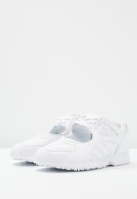 Vita atletiska skor med överdel i mesh, texturerad tyg och gummisula. Funktioner inkluderar snörning och utskurna sidopaneler för andningsförmåga.