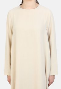 Beige long-sleeve dress with a round neckline, textured fabric, and a relaxed fit. A patterned scarf is tied at the back of the neck.