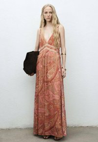 Joven con cabello largo y rubio, que lleva un vestido maxi sin mangas de paisley rosa, sosteniendo una gran bolsa negra contra una pared blanca.
