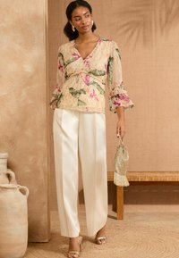 Blusa in chiffon floreale con maniche a volant beige, abbinata a pantaloni in satin crema a vita alta; tiene in mano una piccola borsa in oro con perline.