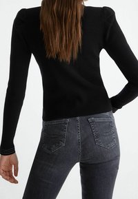 Sort ribstrikket langærmet top med teksturerede skuldre, kombineret med mørkegrå jeans med dekorative syninger på baglommerne.