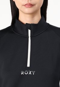 Crna majica na zip s visokim ovratnikom, strukturirana tkanina, bijeli detalj na zipu i bijeli "ROXY" logo na prsima.