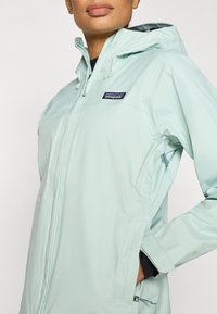 Αδιάβροχο μπουφάν σε ανοιχτό πράσινο μέντας με κουκούλα, διαθέτει εμπρόσθιο φερμουάρ, πλαϊνές τσέπες και λογότυπο στο επάνω αριστερό μέρος του στήθους.