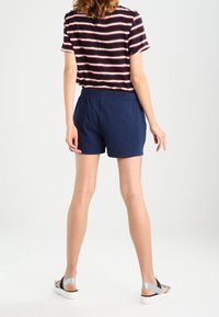 Žena stojící čelem k nám, oblečená v námořnických šortkách, pruhovaném tričku s krátkým rukávem a bílých sandálech se stříbrnými popruhy.