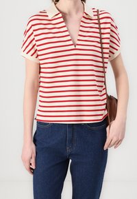 Červeno-bíle pruhovaný polo tričko s krátkým rukávem a hlubokým výstřihem do V, s bledým límcem, spárované s tmavě modrými džíny a hnědou kabelkou přes rameno.