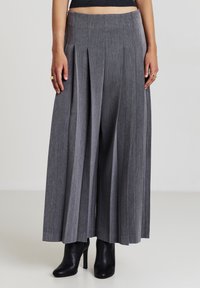 Pantaloni ampi grigi plissettati indossati con stivaletti neri con tacco alto, mostrati su una persona che indossa un top nero corto.