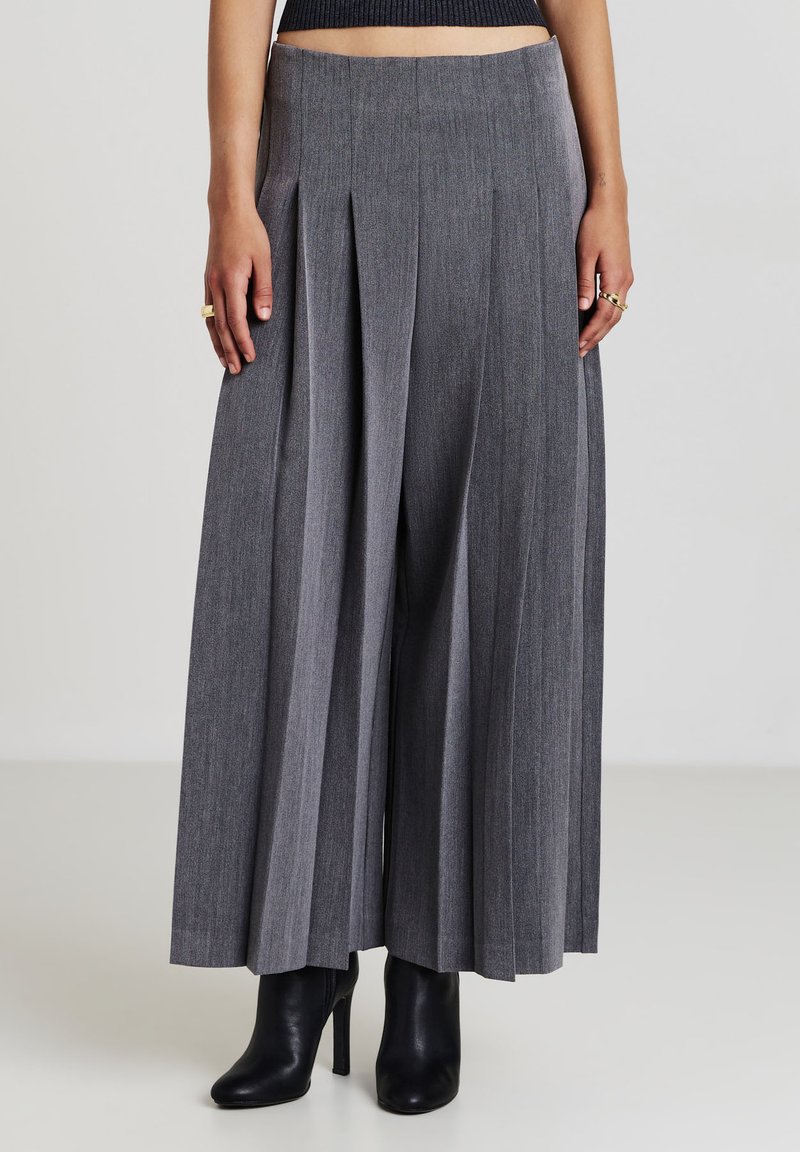 Pantaloni ampi grigi plissettati indossati con stivaletti neri con tacco alto, mostrati su una persona che indossa un top nero corto.