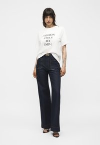 Γυναίκα με σκούρα μαλλιά φοράει λευκό t-shirt με την επιγραφή «FASHION STOLE MY SMILE», σκουρόχρωμο τζιν με ψηλή μέση και φαρδιά πόδια, μαύρα σανδάλια και μαύρη ζώνη.