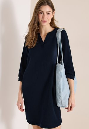 Kobieta w ciemnogranatowej sukience z rękawami do łokci, nosząca na ramieniu jasnoniebieską torbę typu tote, delikatnie uśmiechająca się.