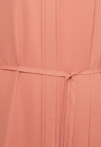 Vestido de tecido cor pêssego com pregas, um cinto atado na cintura e uma textura suave. Sem padrões visíveis ou acessórios.