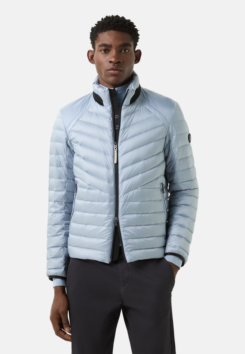 Light blue puffer jacket with a zip front, chevron quilting, high collar, and black accents. Features zippered pockets at the sides.