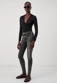 Bodysuit negro de manga larga con escote en V profundo y botones en la parte delantera, combinado con jeans ajustados de talle alto gris y sandalias planas negras.