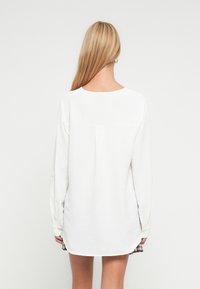 Blouse blanche à manches longues en tissu texturé, avec encolure ronde et pli au dos, associée à une jupe à carreaux.