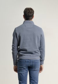 Suéter gris con cuello alto, textura acanalada y mangas ajustadas, combinado con jeans azules que tienen bolsillos traseros con costuras decorativas.