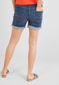 Henkilö, joka on pukeutunut ylös kiinnitettyihin tummansinisiin farkkushortseihin ja raidallisiin liukuviin sandaaleihin, seisoo kädet taskuissa yksinkertaista taustaa vasten.